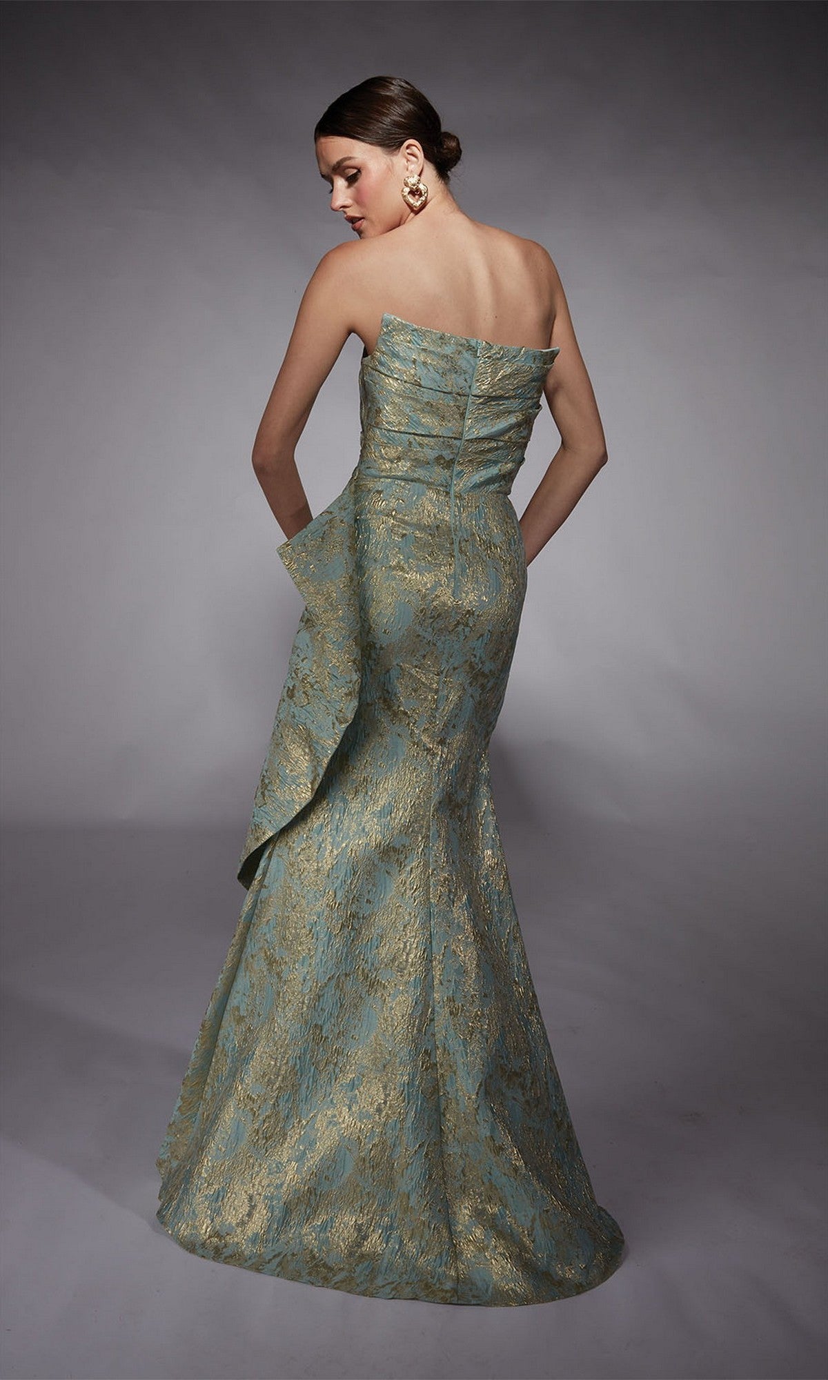 A woman in the Alyce 27750 long formal dress, featuring a strapless gold lace pattern, side drape, metallic jacquard brocade finish, and mermaid skirt, stands with her back to the camera against a neutral background.